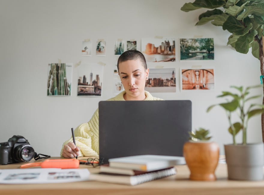 A focused woman designer working on a laptop with a tablet pen in a modern office setup with plants.