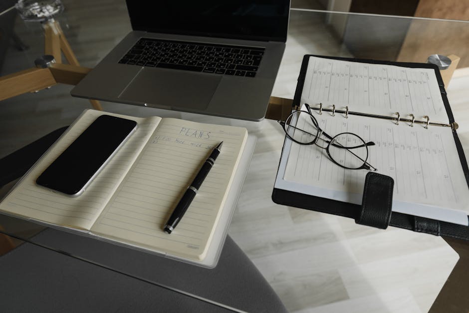 A neatly organized workspace featuring a laptop, smartphone, notebook, pen, and planner on a glass table.